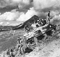 Camouflaged Sherman headed up a steep hill with troopers attending to the tracks