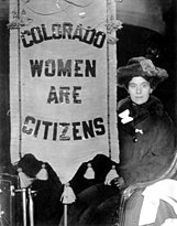 A young woman suffragette sits in a chair near a banner reading "Colorado Women are Citizens." The woman wears a fur coat, hat, and leather gloves. Tassels dangle from the top and bottom of the banner.