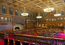 Soudní síň viděná výše, prázdná, ale z jiného úhlu, která ukazuje více a její zdobené stěny, lustry a červený koberec