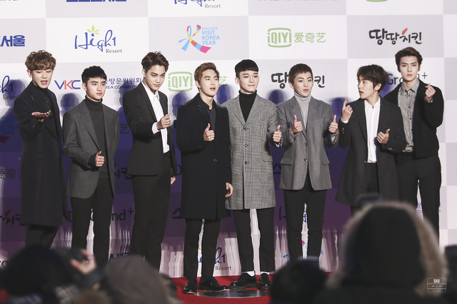 Exo stand in a row on the red carpet, holding their thumbs up and having their photographs taken.