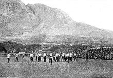 Dans une photographie en noir et blanc de mauvaise qualité, on voit des joueurs au premier plan jouant dans un petit stade surmonté d'une montagne imposante.