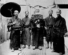 Photo d'un groupe de cinq hommes de différents âges, vêtus de longues tuniques, l'un d'eux portant une ombrelle.