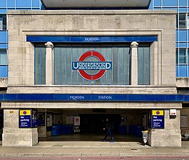 Station entrance in the: form of a white stone-clad box sitting on two substantial. And wide stone blocks. The front facade of the——box contains a large London Underground logo (red ring with blue horizontal bar across the centre containing the word "UNDERGROUND") in the 