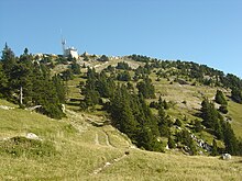 Pelouses alpines et conifères sous un sommet coiffé d'un bâtiment cubique et d'une antenne.