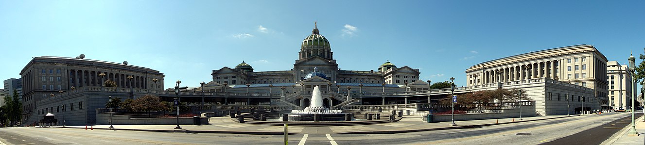 A large plaza with a fountain in front of staircases leading t0 the second level of the plaza. The entire plaza is situated in front of and below a large, domed building. Two large rectangular buildings flank the plaza on either side.