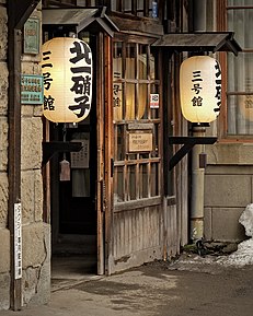 Uma casa cuja fachada possui traços do estilo wabi-sabi, com uma janela de madeira que demonstra a passagem do tempo por estar mais desgastada e lanternas antigas com caligrafia tradicional japonesa