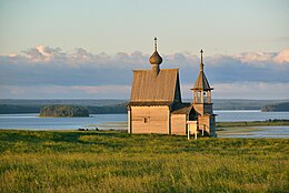 Photographie d'une église en bois d'architecture en bois russe sur une colline avec un lac et des étendues de taïga en arrière-plan.