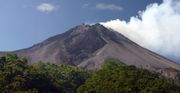 A photograph depicting a blue sky with white clouds at the top, a dark grey volcano in the middle, and green foliage at the bottom.