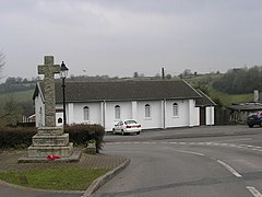 Street scene with stone cross on three tier plinth to the left of the road. In the background is a white walled building.