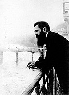 A long-bearded man in his early forties leaning over a railing with a bridge in the background.