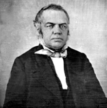 Black and white photo of fair-skinned man, head and shoulders position, hair greying at the temples, wearing a mid-19th century dark jacket and white shirt, loosely tied cravat