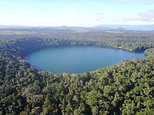 Luftaufnahme des Lake Eacham, umgeben von dichtem tropischem Regenwald, mit der Landnutzung der Atherton Tablelnds im Hintergrund.