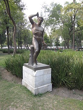 Sculpture of a muscular, nude man in a city park. He stands in contrapposto and prepares to throw a weapon.