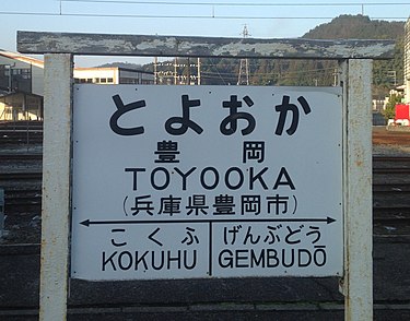 Former Japan National Railways-style board of Toyooka Station. Between the two adjacent stations, "GEMBUDO" follows the Hepburn romanization system, but "KOKUHU" follows the Nihon-shiki/Kunrei-shiki romanization system. Toyooka Station Sign (cropped).jpg