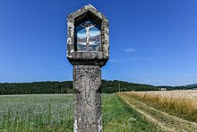 In der Bildmitte befindet sich der Kopf eines Tabernakelpfeilers. Darin wird der gekreuzigte Christus dargestellt. In den Schaft des Pfeilers ist ein Pflugschar eingraviert. Im Bildhintergrund sind grüne Felder und Hügel zu sehen. Rechts neben dem Schaft verläuft ein Weg durch das Feld