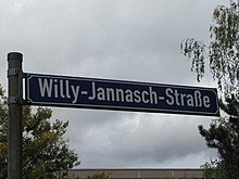 Blaues Straßenschild mit weißer Schrift. Aufschrift: Willy-Jannasch-Straße. Im Hintergrund sind links und rechts Bäume zu sehen. Unten ist das Dach eines Plattenbaus abgebildet.