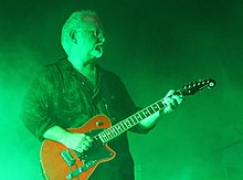 An older man with a gray beard playing a red guitar against a green backlight