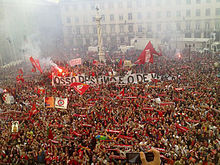 A crowd of Benfica supporters fills the Lisbon City Square. Scarves, flags, banners, and flares can be seen from the City Hall's central balcony.