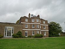 A three-storey brown brick building with a cupola, and a single storey extension on the left, the foreground is a green lawn