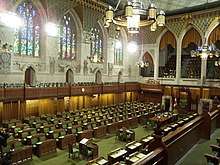 A green-carpeted neo-Gothic-style room with stained-glass windows. There are two banks of chairs facing each other, with three desks in the middle and a gallery off to the side