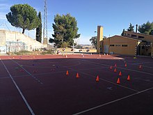 En la fotografía se ve el patio de un colegio. En la cancha de color naranja hay mucho material: conos, cuerdas, picas. En el suelo hay también una serie de líneas que delimitan los diferentes campos. Al fondo un edificio y una portería