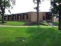 Penn Lake, a beige brick building of one story, sits before a green lawn with punctuated by several tall trees on a sunny summer day.