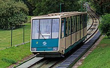 Un funicular verde și bej coboară pe un deal verde.