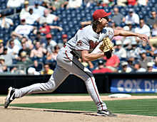 Мужчина в светлой бейсбольной форме и красной кепке бросает мяч.