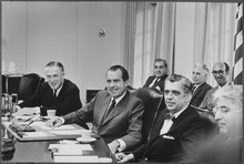 Two middle-aged men sit at the right side of a large tabletop, with others alongside and behind them.