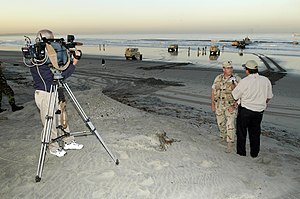 A cameraman at a distance is filming a man interviewing a military officer in fatigues on a beach