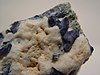 A rough rock showing several intense, dark blue benitoite crystals emerging from white natrolite matrix.