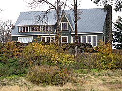 Photograph of a two-story house with a high peak and in a rocky setting with several trees
