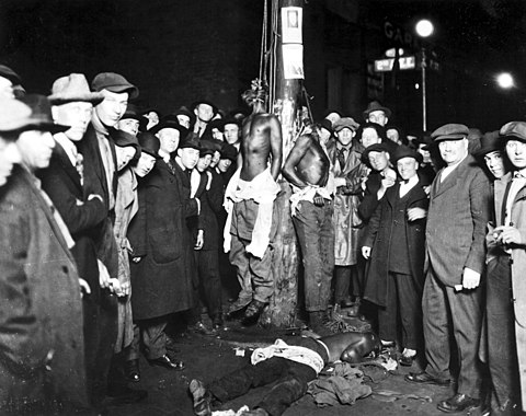 White men pose for a photograph of the 1920 Duluth, Minnesota lynchings. Two of the Black victims are still hanging while the third is on the ground. Lynchings were often public spectacles for the white community to celebrate white supremacy in the U.S., and photos were often sold as postcards.[27]
