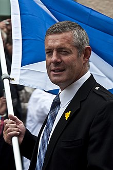 Photographie d'un homme en habillé costume-cravate, Gavin Hastings, tenant dans ses mains le mât du drapeau écossais lors d'une marche.