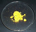 Un poco de polvo de color amarillo canario se asienta, en su mayoría en grumos, en un cristal de reloj de laboratorio.