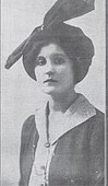 Posed black and white portrain image of a woman in a dark dress and hat seated at a table with papers in front of her and a pen in her hand. A large book is stood upright on the desk and behind the woman are drawers and a fire place.