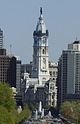 Distant ground-level view of a large building with a white exterior and a tall spire; the spire has a rounded roof and is topped with a black statue.