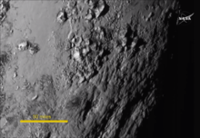 Un petit morceau de la surface de Pluton vue par New Horizons le 14 juillet 2015. La barre jaune qui sert d'échelle représente 80,5 kilomètres (ou 50 miles). Des montagnes d'environ 3 500 m sont visibles et la surface semble récente à la vue de l'absence de cratères[114].