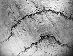 Das Bild ist eine Luftaufnahme einer westlichen Frontlinie im Ersten Weltkrieg. Es sind zwei kurvige Schützengräben als schwarze breite Linie, die horizontal durch das Bild verlaufen, zu erkennen. Außerdem sind Krater von Artilleriegranaten, als kleine Kreise, zu erkennen.