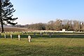 Vista geral do cemitério gramado com suas muitas estelas e cruzes alinhadas.  Uma bandeira francesa hasteada em um mastro.  Ao fundo, árvores e um grande campo arado.