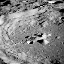 A gray, many-ridged surface from high above. The largest feature is a circular ringed structure with high walled sides and a lower central peak: the entire surface out to the horizon is filled with similar structures that are smaller and overlapping.