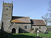 A stone church seen from the south, with a square battlemented tower at the left, then the nave, and a lower chancel at the right. In front of the church is the base of a medieval cross