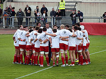 Gracze rugby ubrani w koszulki zbierają się w kółko, trzymając się za ramiona.