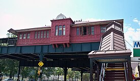 A decorative one-story red building on green steel supports above a street with a red-roofed stair leading up to it. The middle of the building projects slightly forward and its roof is slightly higher than the rest of the building.