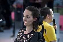 Jeune femme aux cheveux châtains, mi-long assise dans un fauteuil roulant, avec un casque-micro.