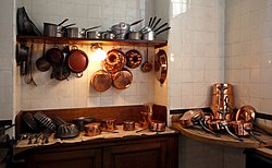 a shelf with kitchen utensils of several kinds stacked upon it, with more utensils hanging from hooks below it, both above two work surfaces with yet further utensils laid out neatly upon them
