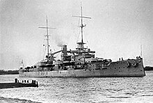 A large battleship lined with guns and equipped with two tall masts sits in harbor.