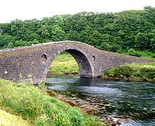 An arched stone bridge crosses a small body of water leading to woodlands in the background