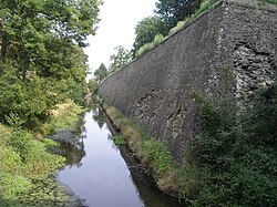 Photo shows a old stone fortress wall about 5 meters tall at right, a wet ditch in the center and trees at left.