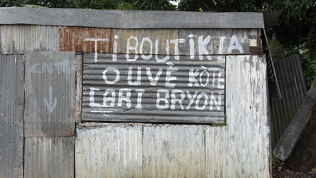 Creole sign in Guadeloupe reading Ti boutik-la ouvè kòté Lari Bryon ("Entrance to the little shop at rue Brion"). Postposition of the definite article
